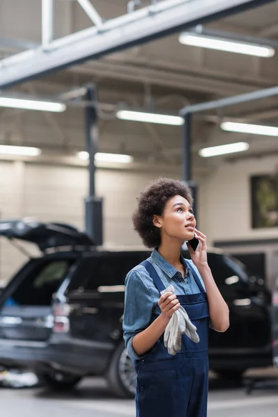 Dreamy young african american mechanic in overalls speaking on smartphone in auto repair service — Stock Photo