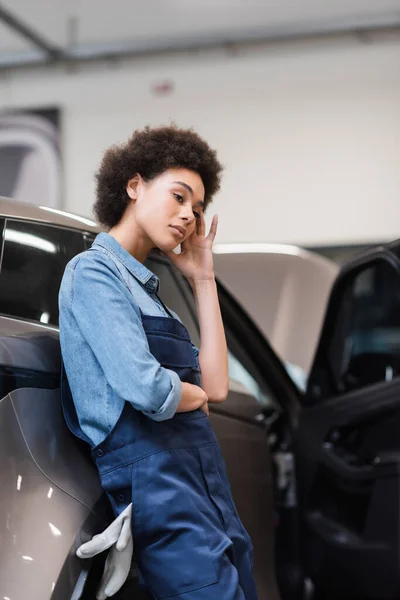 Upset young african american mechanic standing with hand near face in auto repair service — Stock Photo