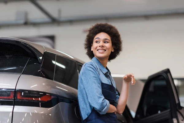 Lächelnder junger afrikanisch-amerikanischer Mechaniker steht mit nahem Auto in Autoreparatur — Stockfoto