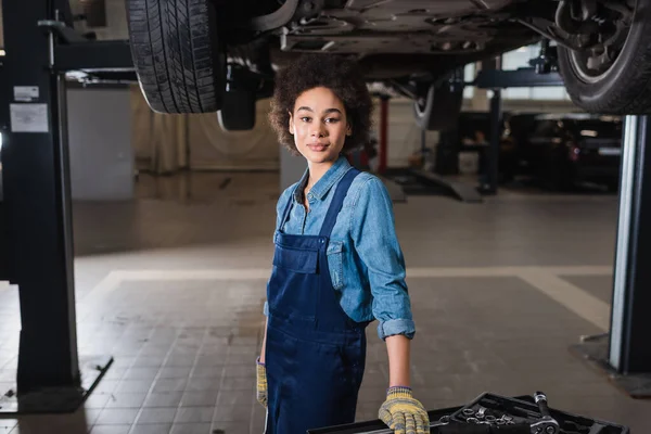 Confident young african american mechanic standing beneath lifted car and looking at camera in garage — Stock Photo