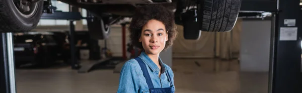 Seguro de sí mismo joven afroamericano mecánico de pie debajo de coche levantado en el garaje, pancarta - foto de stock