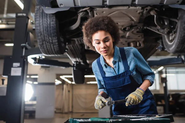 Young african american mechanic standing with wrench beneath lifted car in garage — Stock Photo