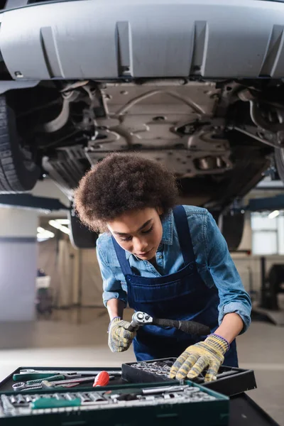 Joven afroamericano mecánico de pie debajo de coche levantado en el garaje - foto de stock