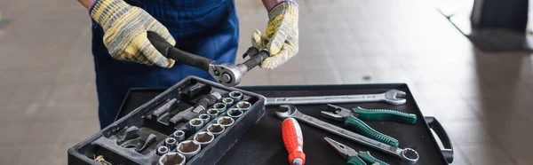 Vue recadrée du mécanicien avec les mains dans les gants tenant la clé près des boîtes à outils dans le garage, bannière — Photo de stock