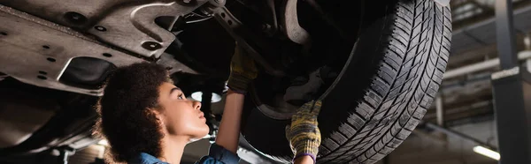 Giovane meccanico afro-americano che ripara ruota di auto sollevata in garage, banner — Foto stock