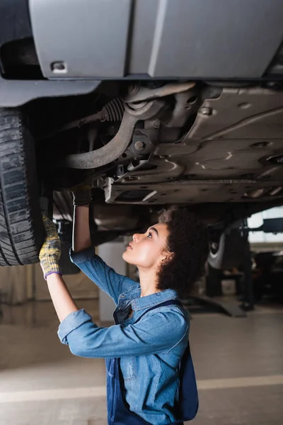 Young african american mechanic repairing bottom of lifted car in garage — Stock Photo