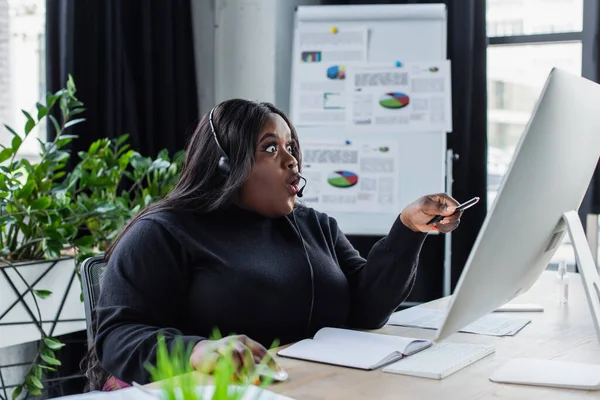 Sorprendido afroamericano más operador de tamaño en auriculares con micrófono apuntando al monitor de computadora en la oficina - foto de stock