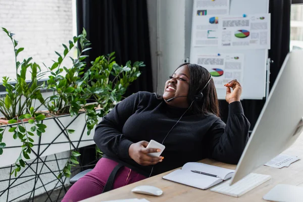 Feliz Africano americano plus size operador em fone de ouvido com microfone falando e segurando smartphone no escritório — Fotografia de Stock