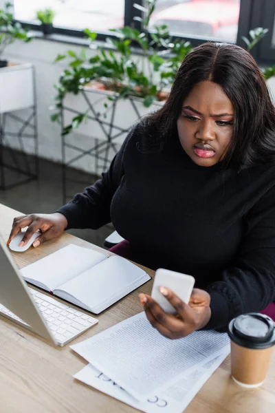 Афроамериканська бізнес-леді плюс розмір дивиться на смартфон біля монітора комп'ютера в офісі — стокове фото