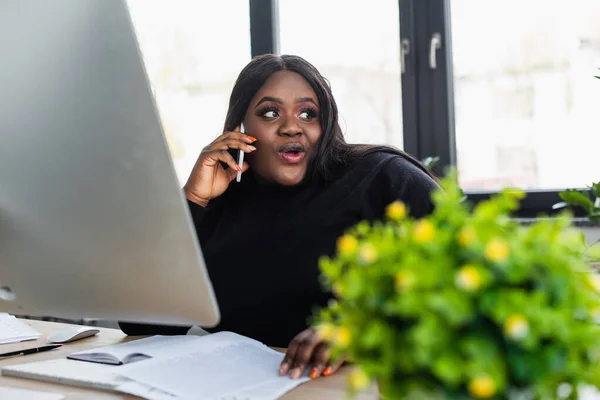Afro-américaine plus taille femme d'affaires parlant sur smartphone près de moniteur d'ordinateur dans le bureau — Photo de stock