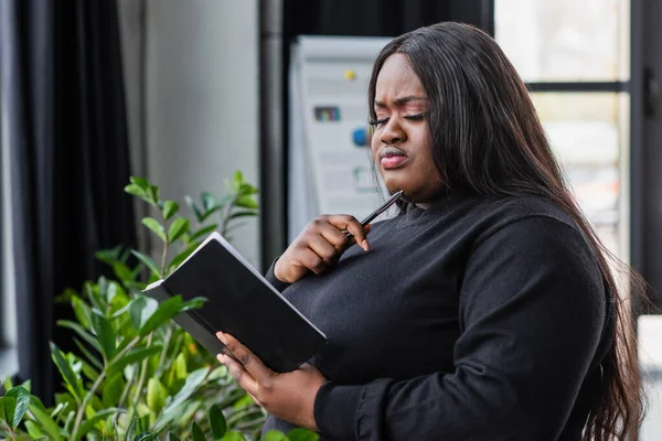 Pensive african american plus size businesswoman holding notebook in office — Stock Photo