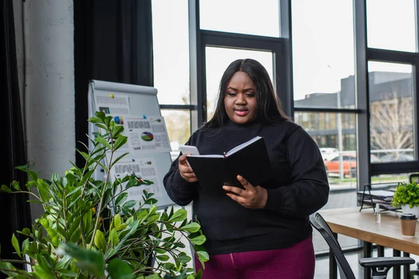 African american plus size businesswoman holding notebook and smartphone in office — Stock Photo