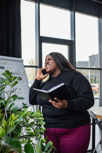 Счастливый африканский американец плюс размер деловая женщина держит ноутбук и говорить на смартфоне в офисе — стоковое фото