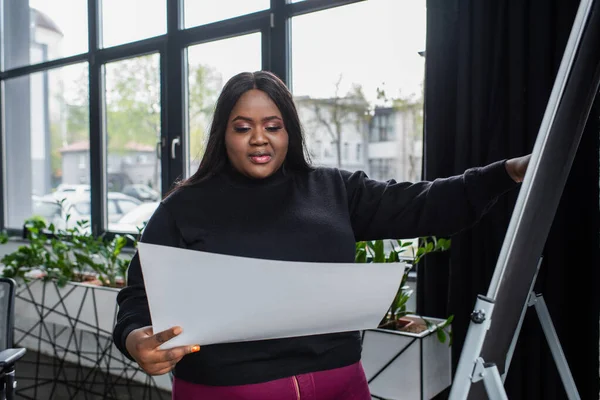 Africano americano más tamaño mujer de negocios sosteniendo papel en blanco cerca de rotafolio - foto de stock