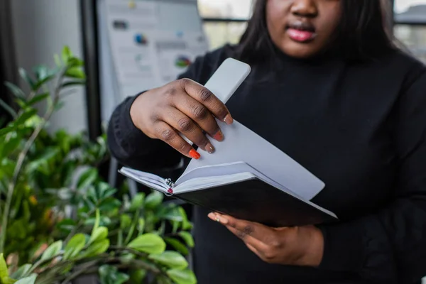 Cropped view of african american plus size woman holding notebook and smartphone in office — Stock Photo