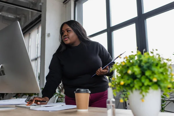 Afroamericana más tamaño mujer de negocios sosteniendo portapapeles y mirando el monitor de la computadora en la oficina - foto de stock