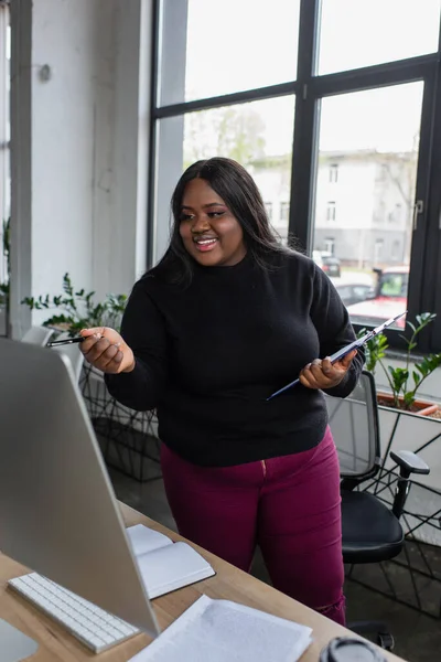 Sonriente afroamericano más tamaño mujer de negocios sosteniendo portapapeles y pluma cerca de monitor de computadora en la oficina - foto de stock