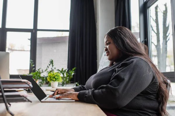 Feliz afro-americano plus size mulher digitando no teclado do laptop no escritório — Fotografia de Stock