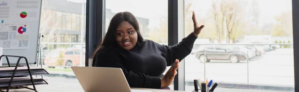 Feliz afro-americano plus size empresária gestos ao ter chamada de vídeo no laptop no escritório, banner — Fotografia de Stock