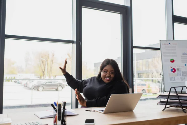 Feliz afro-americano plus size mulher gestos ao ter chamada de vídeo no laptop no escritório — Fotografia de Stock