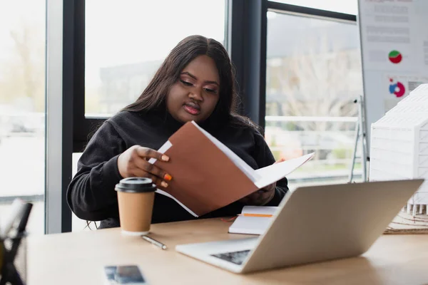 Africano americano más tamaño mujer mirando carpeta cerca de gadgets y taza de papel en escritorio — Stock Photo