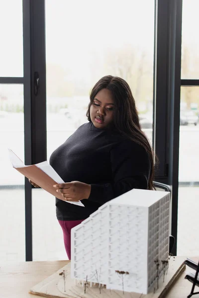 Africano americano más tamaño ingeniero celebración carpeta y mirando modelo de casa - foto de stock