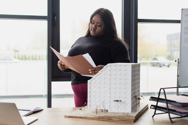 Брюнетка африканский американец плюс размер инженер держит папку рядом дом модели на столе — стоковое фото