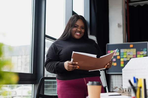 Sonriente afroamericano más tamaño mujer celebración carpeta en la oficina - foto de stock