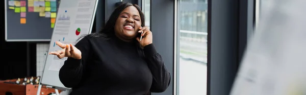 Sorridente afro-americano plus size mulher falando no smartphone no escritório, banner — Fotografia de Stock