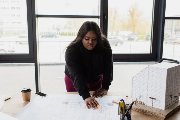 Africano americano más tamaño diseñador mirando plano en escritorio con modelo de casa - foto de stock