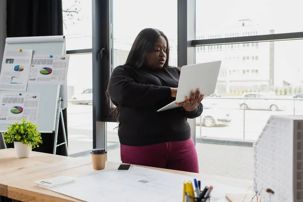 Designer africano americano plus size utilizzando laptop vicino a cianografia sulla scrivania — Foto stock