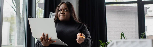 African american plus size woman holding laptop and pointing with pencil, banner — Stock Photo