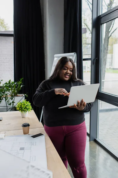 Smiling african american plus size engineer holding laptop near blueprint on desk — Stock Photo