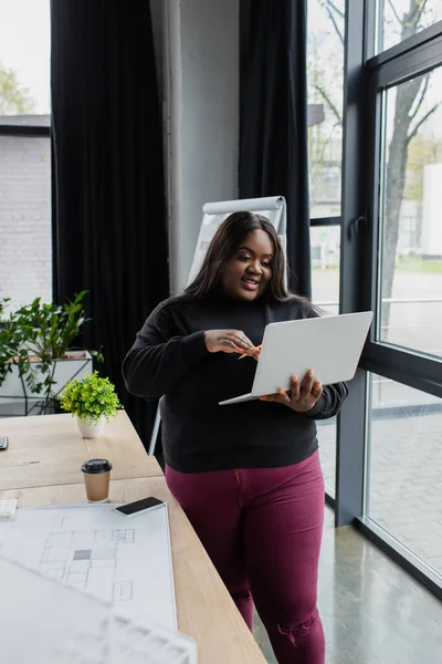 Sorridente Africano americano plus tamanho engenheiro segurando laptop perto de planta na mesa — Fotografia de Stock