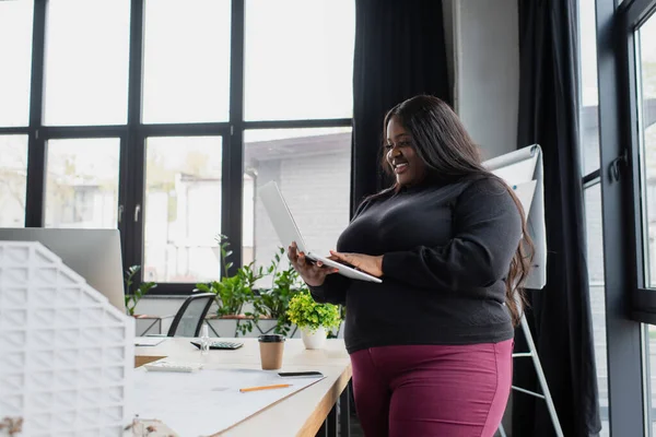Happy african american plus size engineer holding laptop near blueprint and carton house model — Stock Photo