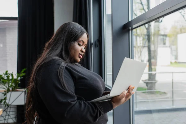 Afrikanisch-amerikanische Plus-Size-Unternehmerin hält Laptop neben Fenster im Büro — Stockfoto