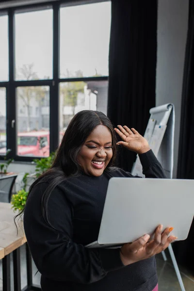 Joyeux afro-américain plus taille femme d'affaires tenant un ordinateur portable et agitant la main tout en ayant appel vidéo — Photo de stock