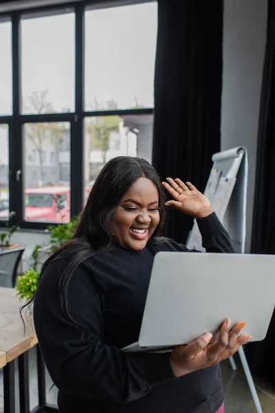 Alegre Africano americano plus tamanho empresária segurando laptop e mão acenando enquanto tem chamada de vídeo — Fotografia de Stock