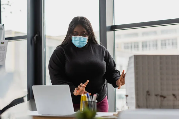 Afrikanisch-amerikanische Plus-Size-Unternehmerin in medizinischer Maske gestikuliert bei Videoanruf auf Laptop im Büro — Stockfoto