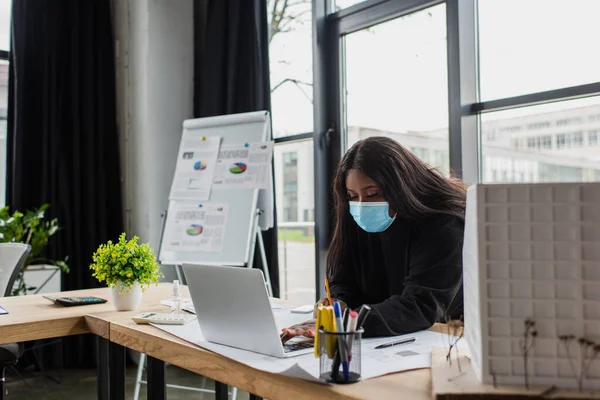 Afroamericano plus size ingegnere in maschera medica utilizzando laptop vicino a cianografia e modello di casa in ufficio — Foto stock