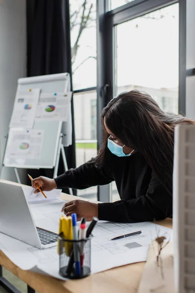 Afrikanisch-amerikanischer Plus-Size-Ingenieur in medizinischer Maske schaut sich Bauplan in der Nähe von Laptop und Hausmodell im Büro an — Stockfoto