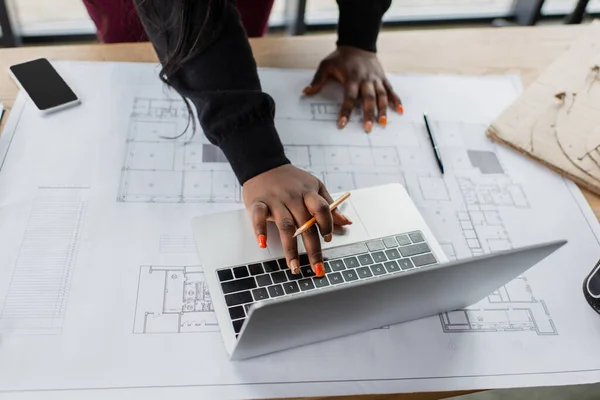 Cropped view of african american female engineer using laptop near blueprint on desk — Stock Photo