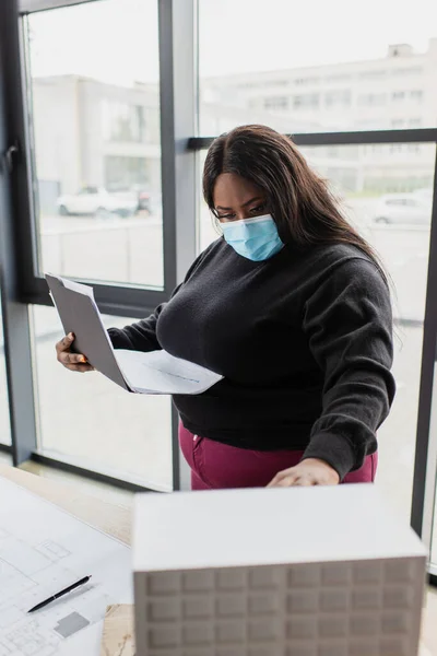 Africano americano más tamaño ingeniero en médico máscara celebración carpeta cerca borrosa casa modelo en oficina - foto de stock