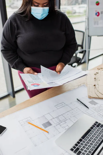 African american plus size engineer in medical mask holding charts and graphs near blueprint — Photo de stock