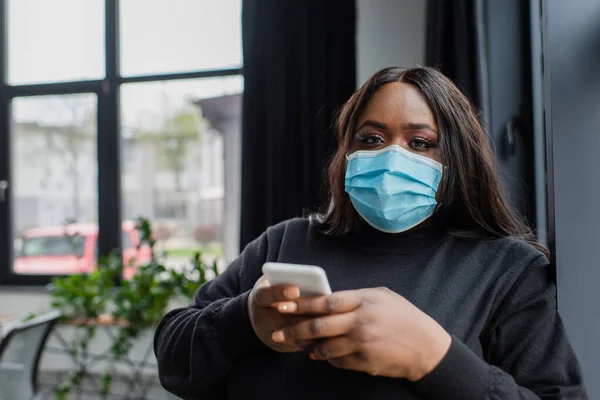 Afro-américaine plus taille femme d'affaires en masque médical en utilisant smartphone dans le bureau — Photo de stock