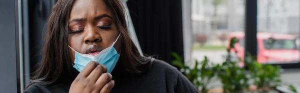Africano americano más tamaño mujer de negocios quitándose la máscara médica mientras se siente mal en la oficina, pancarta - foto de stock