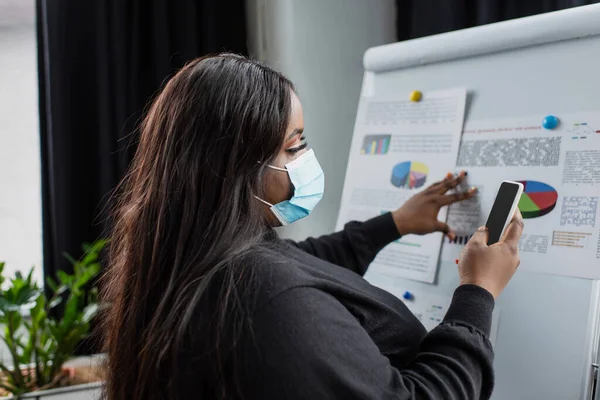 Африканский американец плюс размер деловая женщина в медицинской маске с помощью смартфона рядом флип-чарт с диаграммами и графиками — стоковое фото