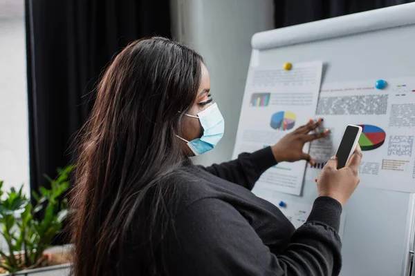 Africano americano plus tamanho empresária em máscara médica usando smartphone perto de flip chart com gráficos e gráficos — Fotografia de Stock