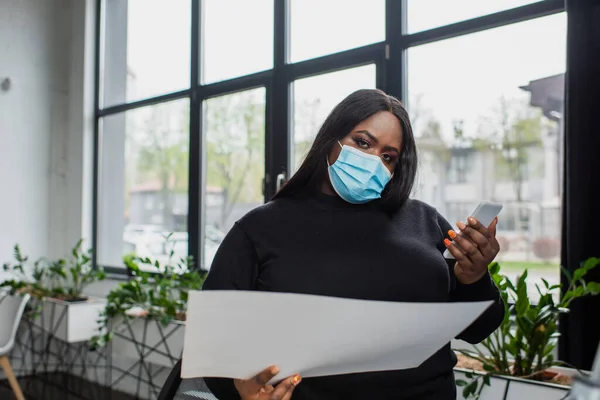 Afroamericano plus size donna d'affari in maschera medica utilizzando smartphone mentre in possesso di documento — Foto stock