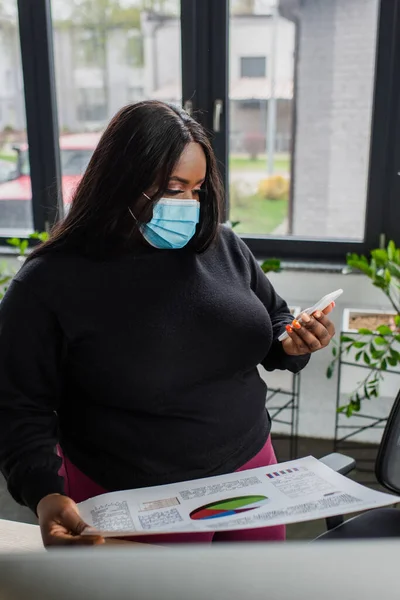 Africano americano más tamaño mujer de negocios en máscara médica celebración de teléfono inteligente y papel con gráficos y gráficos - foto de stock